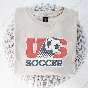 May include: A light beige crew neck t-shirt featuring a graphic design with the red text US and a soccer ball illustration with motion lines. The word SOCCER is printed in blue below the graphic. The design has a distressed, weathered texture.