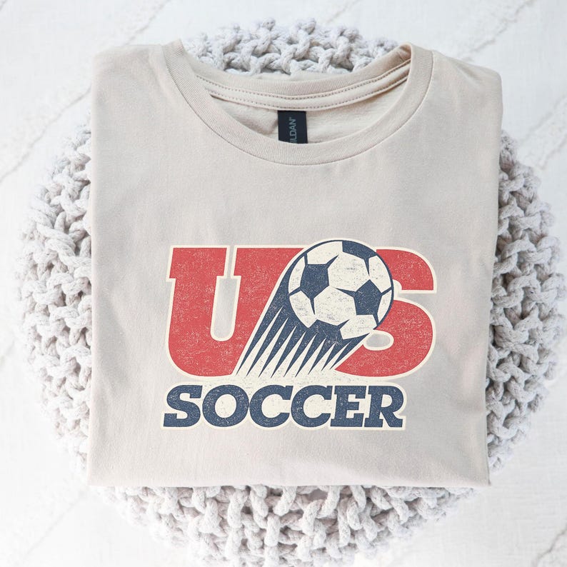 May include: A light beige crew neck t-shirt featuring a graphic design with the red text US and a soccer ball illustration with motion lines. The word SOCCER is printed in blue below the graphic. The design has a distressed, weathered texture.