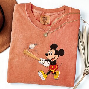 May include: A coral-coloured t-shirt featuring a cartoon of Mickey Mouse swinging a baseball bat at a baseball. The shirt has a Comfort Colors tag and a gold necklace. The shirt is a small size.