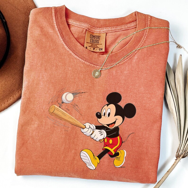 May include: A coral-coloured t-shirt featuring a cartoon of Mickey Mouse swinging a baseball bat at a baseball. The shirt has a Comfort Colors tag and a gold necklace. The shirt is a small size.