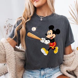 May include: Dark grey t-shirt featuring a cartoon Mickey Mouse character swinging a baseball bat at a baseball. Mickey Mouse is wearing yellow shoes, red shorts, and white gloves. The baseball is white with red stitching.