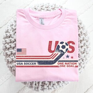 May include: A light pink t-shirt featuring a graphic with the text US, USA SOCCER, and ONE NATION ONE GOAL. The design includes a soccer ball, a small American flag, and horizontal red, white, and blue stripes with a distressed, vintage-style texture.