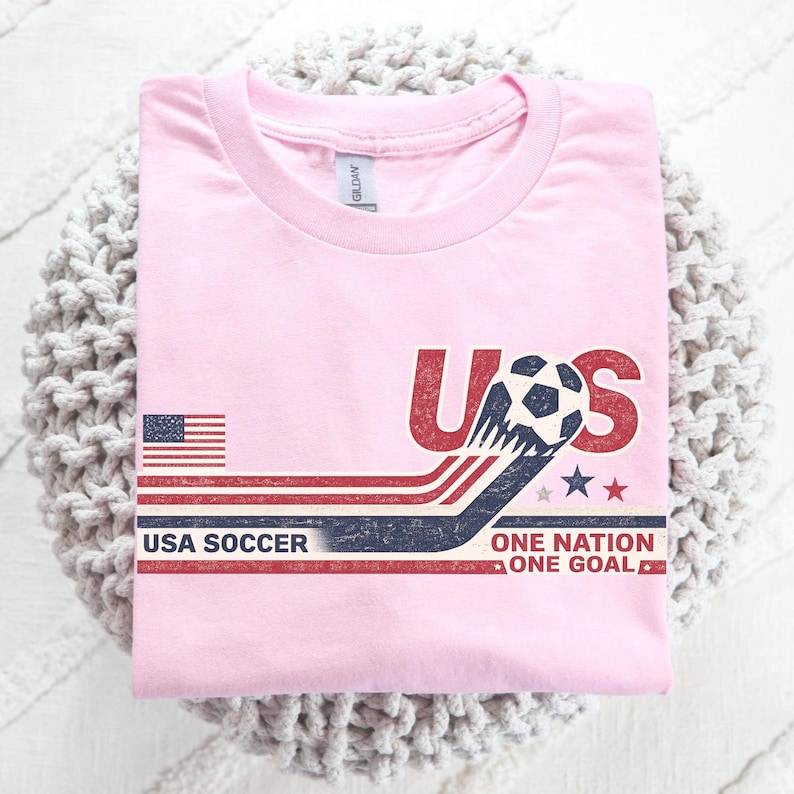May include: A light pink t-shirt featuring a graphic with the text US, USA SOCCER, and ONE NATION ONE GOAL. The design includes a soccer ball, a small American flag, and horizontal red, white, and blue stripes with a distressed, vintage-style texture.