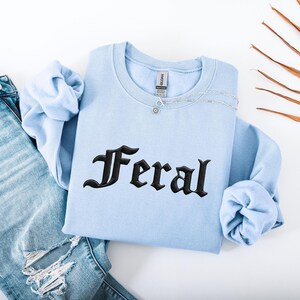May include: A light blue sweatshirt with the word "Feral" embroidered in black gothic lettering. A silver necklace with a small pendant rests on the sweatshirt. The sleeves are folded, and a pair of blue jeans is partially visible.