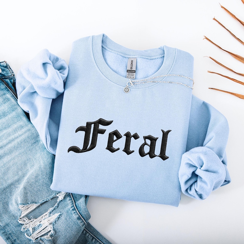May include: A light blue sweatshirt with the word "Feral" embroidered in black gothic lettering. A silver necklace with a small pendant rests on the sweatshirt. The sleeves are folded, and a pair of blue jeans is partially visible.