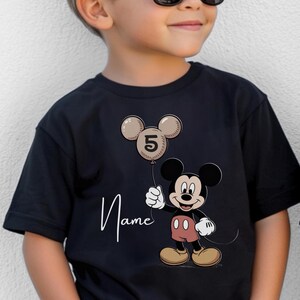 May include: Black children's t-shirt with a Mickey Mouse cartoon holding a beige balloon with the number 5. The word "Name" is written in cursive below the balloon. The shirt is black.
