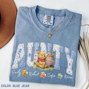 Peut inclure: T-shirt bleu jean Comfort Colors avec "AUNTY" en lettres patchwork. Avec Winnie l'Ourson, un pot de miel et des abeilles. Les noms "Emily", "Robert", "Sofia" et "Karen" sont en dessous. Un collier en or est drapé sur le t-shirt.