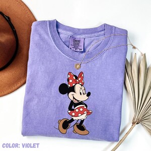 May include: Violet Comfort Colors t-shirt featuring a Minnie Mouse graphic. The cartoon character is wearing a red polka dot bow and skirt. A gold necklace is draped over the shirt. The text "COLOR: VIOLET" is at the bottom.