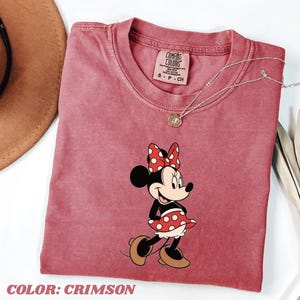 May include: Crimson-colored t-shirt featuring a Minnie Mouse graphic. The cartoon character is depicted in a red and white polka-dot dress and bow. The shirt has a crew neck and a small tag at the neckline. A silver necklace with a pendant is also visible.