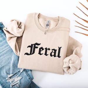 May include: A beige sweatshirt with the word "Feral" embroidered in black gothic lettering. The sweatshirt is folded, with a silver necklace draped across it. Blue jeans are visible in the background.