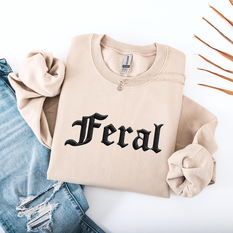 May include: A beige sweatshirt with the word "Feral" embroidered in black gothic lettering. The sweatshirt is folded, with a silver necklace draped across it. Blue jeans are visible in the background.