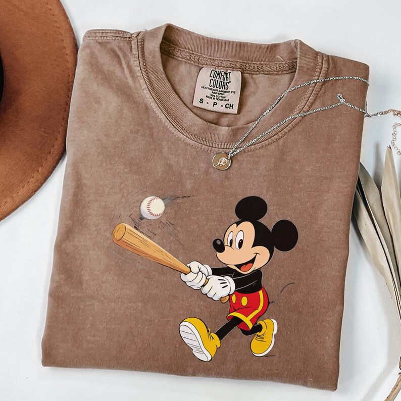 May include: Brown Comfort Colors t-shirt with a Mickey Mouse baseball graphic. The shirt has a crew neck and short sleeves. A silver necklace with a pendant is on the shirt. The shirt is a small size.