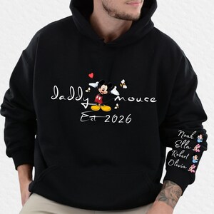 Pode incluir: Moletom preto com a inscri&ccedil;&atilde;o branca "Daddy Mouse Est 2026" e um desenho animado do Mickey Mouse. A manga direita tem nomes e pequenas figuras de desenhos animados.