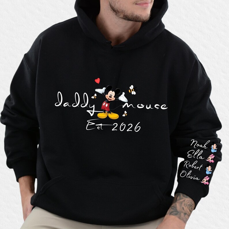 Pode incluir: Moletom preto com a inscri&ccedil;&atilde;o branca "Daddy Mouse Est 2026" e um desenho animado do Mickey Mouse. A manga direita tem nomes e pequenas figuras de desenhos animados.