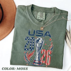K&ouml;nnte beinhalten: Ein moosgr&uuml;nes Kurzarm-T-Shirt mit einem Grafikdruck, der den Text USA 2026, einen Fu&szlig;ballpokal sowie rote, wei&szlig;e und blaue Sterne und Streifen zeigt. Das T-Shirt hat einen Rundhalsausschnitt und ein Comfort Colors-Etikett am Innenhals.