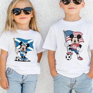 May include: Two white t-shirts featuring cartoon characters. The left shirt shows Minnie Mouse holding a Scottish flag and a soccer ball. The right shirt shows Mickey Mouse wearing a red USA shirt, holding an American flag, and standing on a soccer ball.