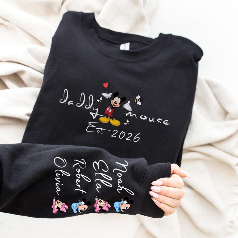 Pode incluir: Camisola preta com as palavras "Daddy mouse" e "Est 2026" em escrita branca. Uma imagem do Mickey Mouse est&aacute; no centro. A manga exibe os nomes Olivia, Robert, Ella e Noah, com imagens do Mickey Mouse.