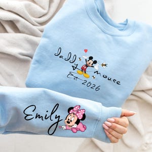 Pode incluir: Camisola azul clara com um design do Mickey Mouse e o texto "Daddy Mouse Est 2026". A manga exibe o nome "Emily" e um gr&aacute;fico da Minnie Mouse. O design inclui abelhas e um cora&ccedil;&atilde;o.