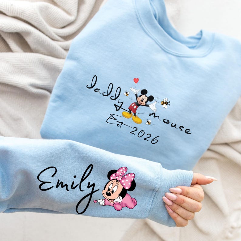 Pode incluir: Camisola azul clara com um design do Mickey Mouse e o texto "Daddy Mouse Est 2026". A manga exibe o nome "Emily" e um gr&aacute;fico da Minnie Mouse. O design inclui abelhas e um cora&ccedil;&atilde;o.