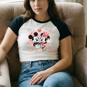 May include: White and black raglan crop top featuring a graphic of Mickey and Minnie Mouse inside a pink heart. The design includes hearts, fireworks, and a gift. The shirt is worn with blue jeans.