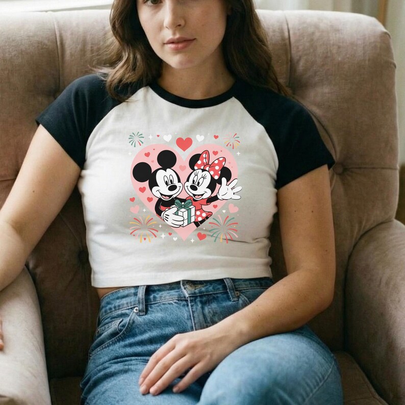 May include: White and black raglan crop top featuring a graphic of Mickey and Minnie Mouse inside a pink heart. The design includes hearts, fireworks, and a gift. The shirt is worn with blue jeans.