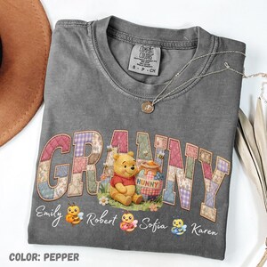 May include: A grey t-shirt with the word "GRANNY" in a patchwork design, featuring Winnie the Pooh and a "HUNNY" pot. Names and bee graphics are below. A silver necklace and a brown hat are visible. The shirt is pepper-colored.