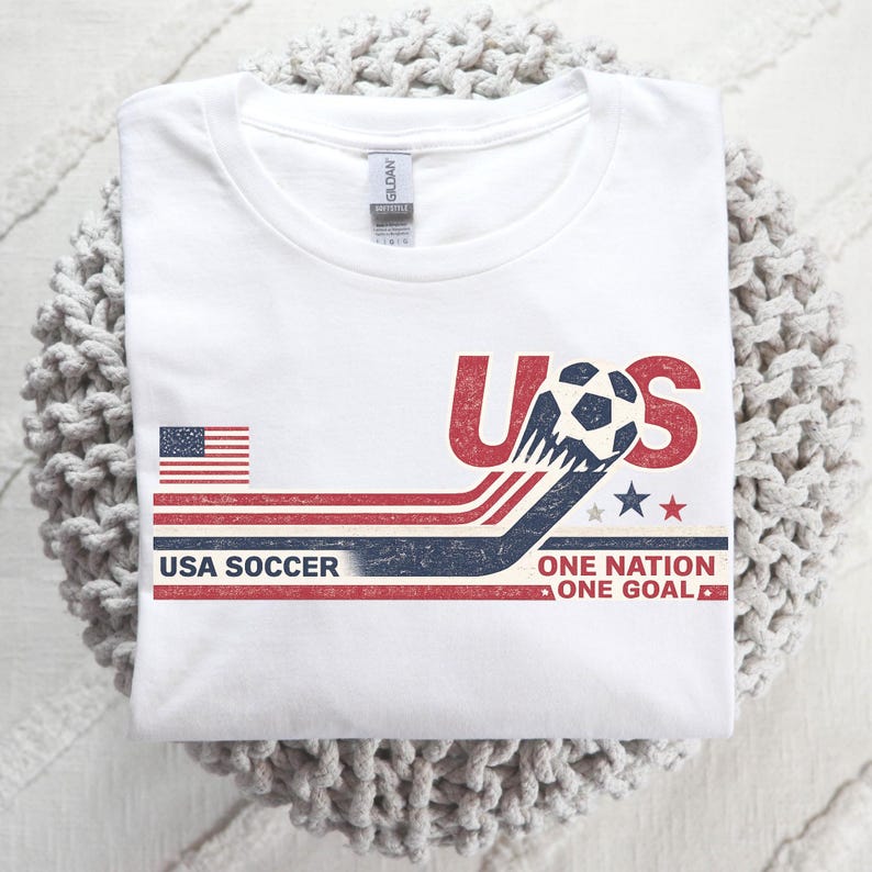 May include: A white short-sleeve t-shirt featuring a graphic with the text USA, USA SOCCER, and ONE NATION ONE GOAL. The design includes a small American flag, a soccer ball, and red, white, and blue stripes with a distressed, vintage-style texture.