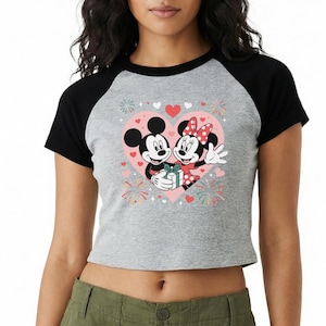 May include: A gray raglan crop top featuring Mickey and Minnie Mouse embracing inside a pink heart. The graphic includes hearts, fireworks, and a gift. The sleeves are black.