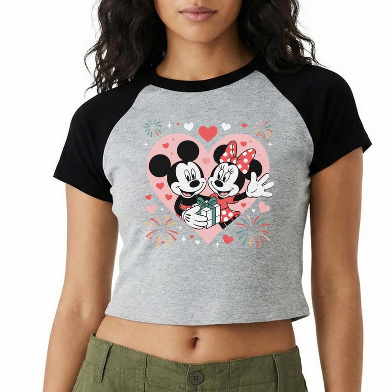 May include: A gray raglan crop top featuring Mickey and Minnie Mouse embracing inside a pink heart. The graphic includes hearts, fireworks, and a gift. The sleeves are black.