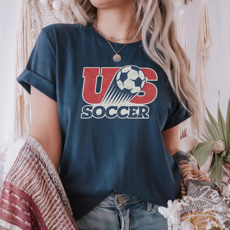 May include: A dark blue short-sleeved t-shirt featuring a graphic design with the red text US and a black and white soccer ball. The word SOCCER is printed in white below the graphic. The design has a distressed, weathered texture throughout.