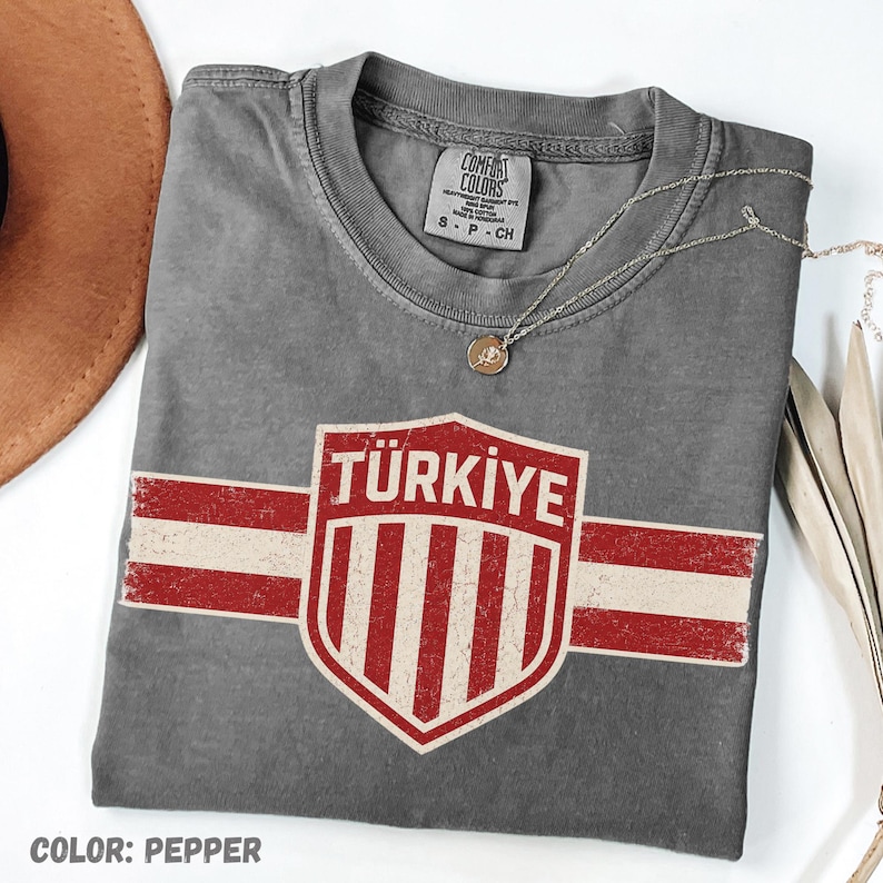 May include: A pepper-colored t-shirt featuring a distressed graphic of a shield with the text T&Uuml;RKIYE above vertical red and white stripes. The shirt has a crew neckline and a Comfort Colors brand label inside the collar, size S-P-CH.