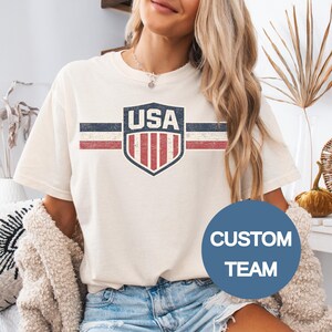 May include: A cream-colored short-sleeve t-shirt featuring a distressed graphic of a shield with the text USA above vertical red and white stripes, flanked by horizontal blue, white, and red lines. A blue circular overlay displays the text CUSTOM TEAM.