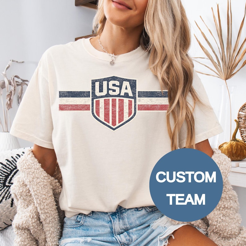 May include: A cream-colored short-sleeve t-shirt featuring a distressed graphic of a shield with the text USA above vertical red and white stripes, flanked by horizontal blue, white, and red lines. A blue circular overlay displays the text CUSTOM TEAM.