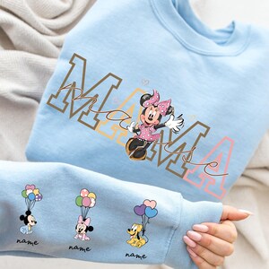 May include: Light blue sweatshirt featuring a Minnie Mouse graphic with the word "MAMA" in brown and pink. The sleeve displays Mickey, Minnie, and Pluto with balloons. Each character has a space for a name. A fun, colorful design.