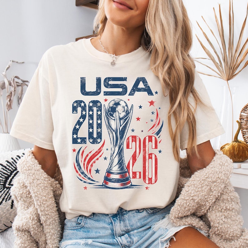 K&ouml;nnte beinhalten: Ein cremefarbenes Kurzarm-T-Shirt mit einem Grafikdruck und dem Text USA 2026. Das Design zeigt eine Fu&szlig;ball-Troph&auml;en-Illustration, rote und blaue Sterne sowie rote und blaue geschwungene Linien in einer abgenutzten, vintage-artigen Textur.