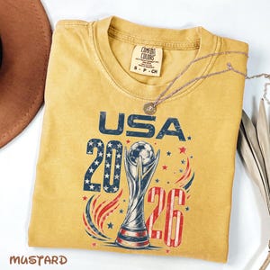 K&ouml;nnte beinhalten: Ein senfgelbes T-Shirt mit einem Grafikdruck, der den Text USA 2026, eine Fu&szlig;ballpokal-Illustration sowie rote, wei&szlig;e und blaue Stern- und Wirbelakzente zeigt. Das Etikett der Marke Comfort Colors befindet sich im Kragenbereich.