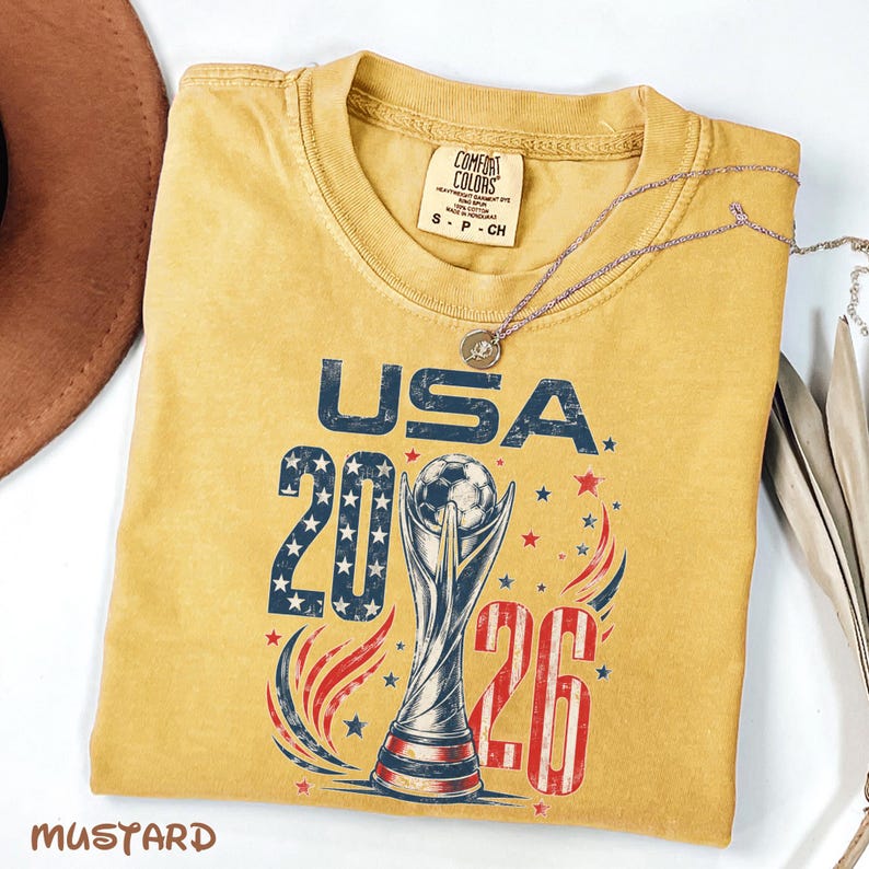 K&ouml;nnte beinhalten: Ein senfgelbes T-Shirt mit einem Grafikdruck, der den Text USA 2026, eine Fu&szlig;ballpokal-Illustration sowie rote, wei&szlig;e und blaue Stern- und Wirbelakzente zeigt. Das Etikett der Marke Comfort Colors befindet sich im Kragenbereich.