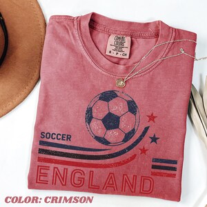 May include: A crimson short-sleeve t-shirt featuring a distressed graphic of a soccer ball, three stars, and the text SOCCER ENGLAND. The shirt has a crew neckline and a Comfort Colors brand label inside the collar, with a thin gold-tone necklace.