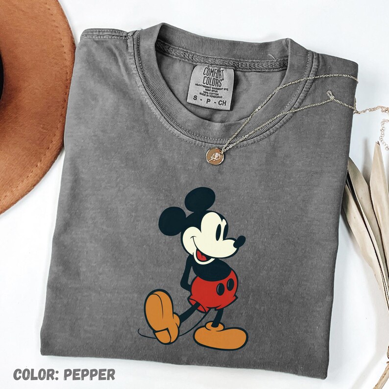 May include: A gray Comfort Colors t-shirt featuring a graphic of Mickey Mouse in red shorts and yellow shoes. The shirt has a crew neck and short sleeves. A silver necklace with a small pendant is draped across the shirt. The text "COLOR: PEPPER" is at the bottom.