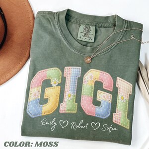May include: Moss green t-shirt with the name "GIGI" in a patchwork design with daisy accents. The shirt also features the names "Emily", "Robert", and "Sofia" below the main text. A gold necklace rests on the shirt.