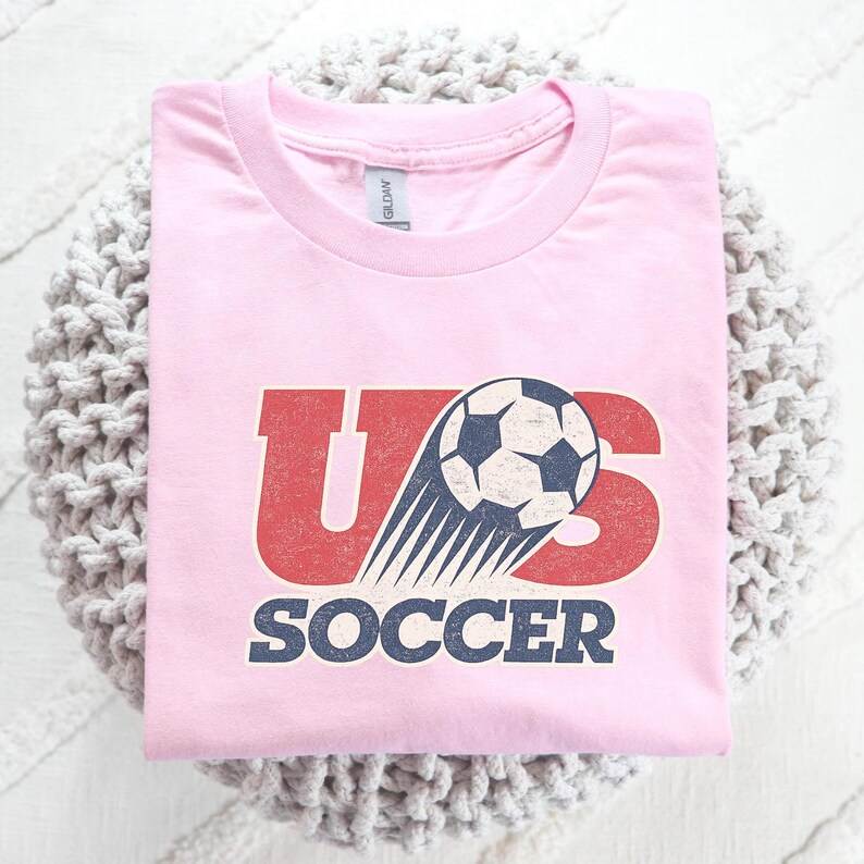 May include: A light pink short-sleeved t-shirt featuring a graphic print with the red letters US, a black and white soccer ball with motion lines, and the word SOCCER in dark blue text below. The design has a distressed, weathered appearance.