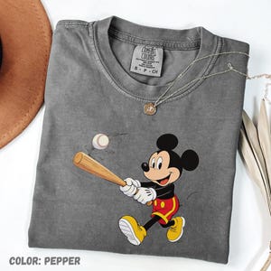 May include: A grey Comfort Colors t-shirt with a cartoon Mickey Mouse playing baseball. Mickey is depicted in red shorts, yellow shoes, and white gloves, swinging a bat at a baseball. The shirt has a crew neck and short sleeves. The text "COLOR: PEPPER" is at the bottom.