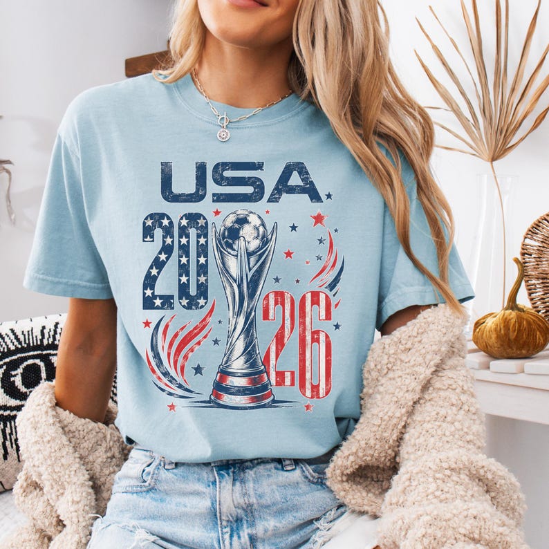 K&ouml;nnte beinhalten: Ein hellblaues T-Shirt mit einem Grafikdruck, der den Text USA 2026, eine Fu&szlig;ballpokal-Illustration sowie rote, wei&szlig;e und blaue Stern- und Wirbelakzente zeigt. Das Design weist eine abgenutzte, vintage-inspirierte Textur im gesamten Druck auf.