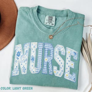 Puede incluir: Camiseta verde claro con la palabra "NURSE" en un diseño de patchwork con estampados florales, cuadros y lunares en azul y blanco. Un collar dorado con un colgante está drapeado sobre la camisa.