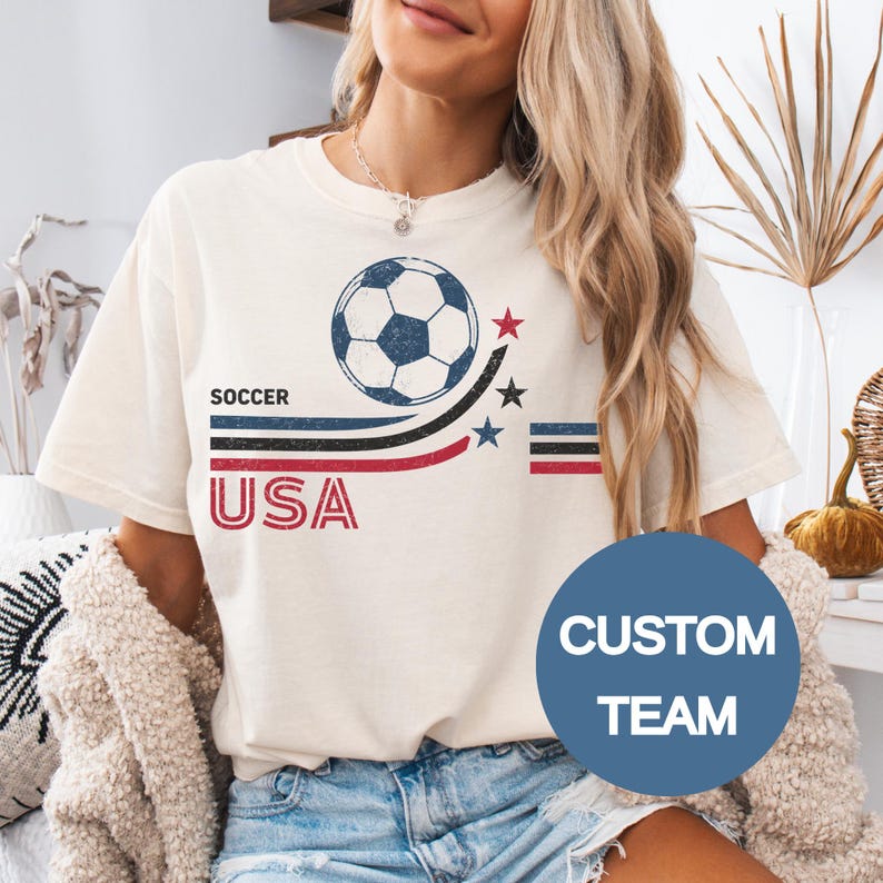 May include: A cream-colored short-sleeve t-shirt featuring a graphic with a soccer ball, the text SOCCER and USA, and three horizontal stripes in black, blue, and red. The design includes three small stars in red, black, and blue colors.