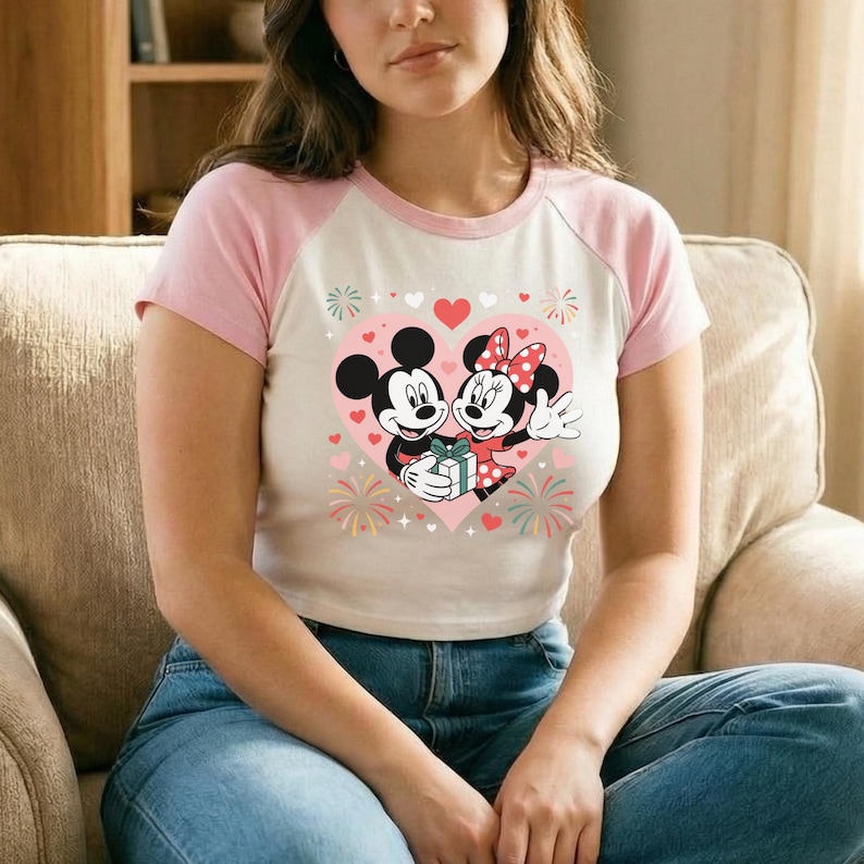 May include: A white and pink raglan t-shirt featuring a graphic of Mickey and Minnie Mouse embracing inside a pink heart. The design includes hearts, fireworks, and a gift. The shirt is worn by a person.