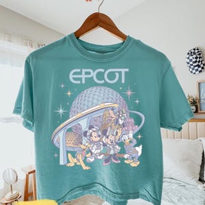 May include: A teal short-sleeve t-shirt featuring a graphic print of Epcot with the Spaceship Earth structure, a monorail, and Disney characters including Mickey, Minnie, Donald, Pluto, and Goofy, with star accents and the text EPCOT.