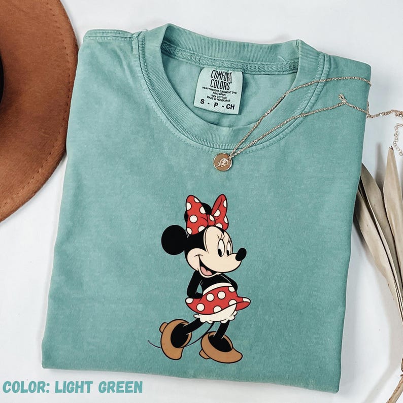 May include: Light green t-shirt featuring a Minnie Mouse graphic. The cartoon character is wearing a red polka dot bow and skirt. The shirt has a crew neck and short sleeves. A gold necklace is draped across the shirt.