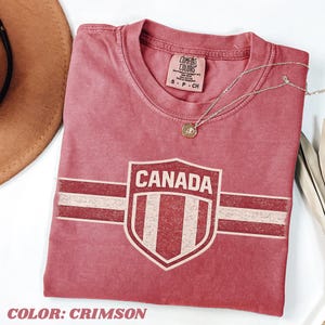 May include: A crimson short-sleeved t-shirt featuring a distressed graphic of a shield with the text CANADA and horizontal stripes. The shirt has a crew neckline and a Comfort Colors brand label on the inside collar. A thin gold chain necklace rests on top.