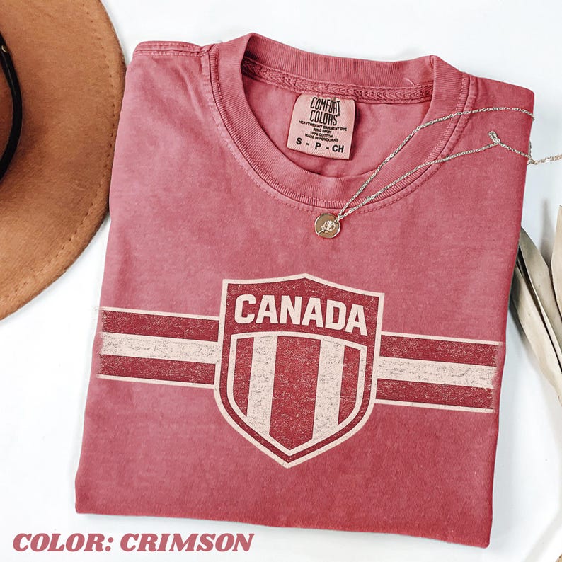 May include: A crimson short-sleeved t-shirt featuring a distressed graphic of a shield with the text CANADA and horizontal stripes. The shirt has a crew neckline and a Comfort Colors brand label on the inside collar. A thin gold chain necklace rests on top.